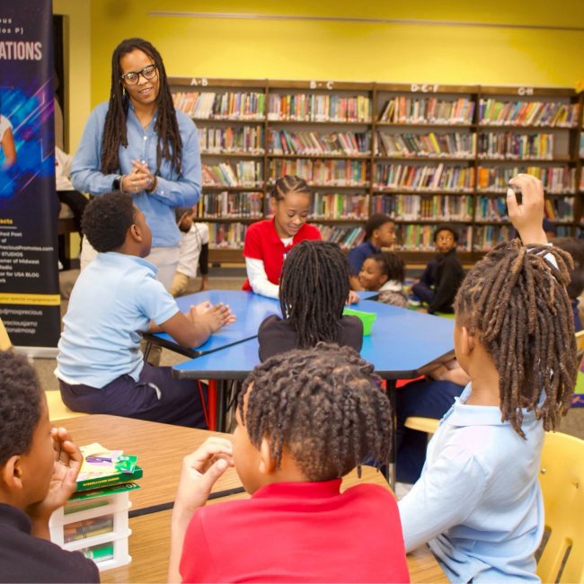 Students seated in a school library raise their hands during an interactive author presentation.