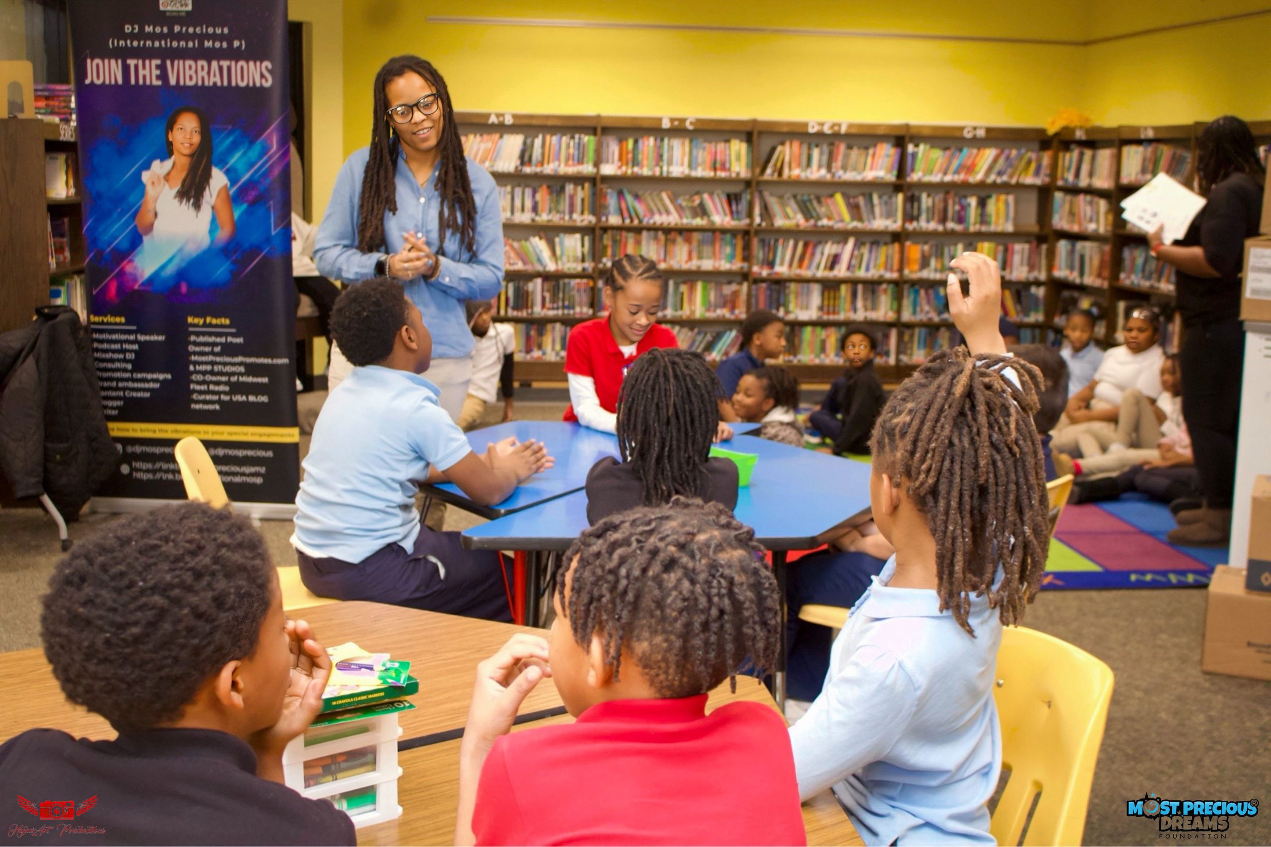 Students seated in a school library raise their hands during an interactive author presentation.