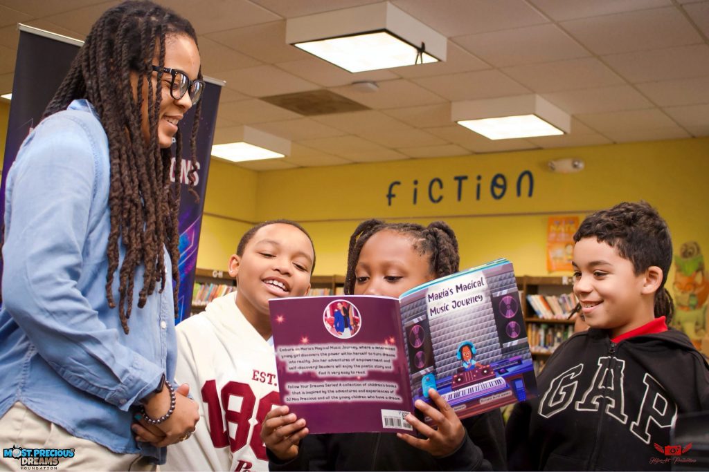 Three smiling students read Maria's Magical Music Journey together while the author watches in a school library.