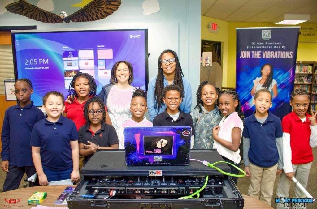 Students and educators pose with Most Precious Dreams Foundation founder Mahogany Henderson behind DJ equipment during a youth literacy and creative arts event.